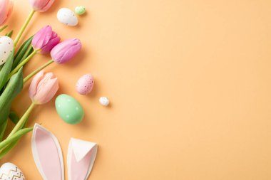 Festivalsel Pasteller: Üst manzara canlı yumurtalar, neşeli tavşan kulakları ve yumuşak turuncu arka planda laleler sergiliyor. Açık alan kişiselleştirilmiş ileti veya reklamları davet eder, Paskalya Nisan ambiyansı özetlenir