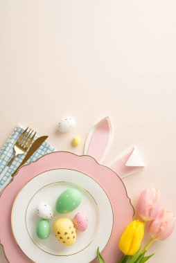 Paskalya sofrası kurma vizyonu. Üst görünüm, canlı yumurtalar, tavşan kulakları, peçete, sofra takımı ve bej arka planda bej renkli lalelerle dolu bir tabağın dikey perspektifi.