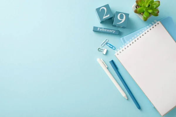 End-of-month workplace scene: overhead shot of notepads, pens, stationery, a potted plant, and a calendar showing February 29th, arranged on a soft blue backdrop with space for text