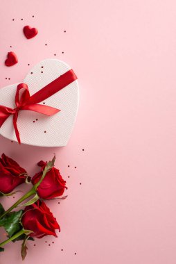Sevgililer Günü için ideal bir kurgu. Zarif güllerin dikey görüntüsü, şık kalp şeklinde hediye, süslemeler ve simler yumuşak pembe bir arka planda, mesajınız veya ilanınız için yer bırakarak.