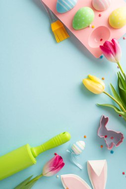 Paskalya pastası hazırlığı. Aletleri olan bir fırın düzeneğinin dikey üst görüntüsü, sevimli kurabiye bıçağı, tutacakta yumurtalar, şeker parçacıkları, tavşan kulakları ve laleler. Pastel mavi arkaplan, metin için açık