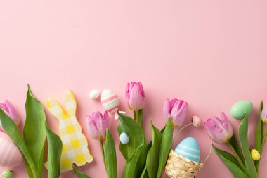 Canlı yumurtalar, sevimli bir tavşan, küçük bir sepet ve lalelerle şenlik düzenlemesi. Pastel pembe bir yüzeyin üst görüntüsü, mesajınız veya reklamınız için hoş bir alan sunuyor.