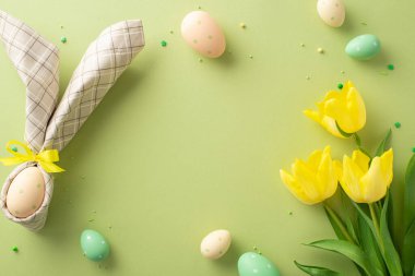 Neşeli Paskalya sahnesi. Tavşan kulaklı peçeteyle süslenmiş bir masa, göz kamaştırıcı sarı laleler, pastel yeşil arka planda şeker serpiştirilmiş. Metin yerleştirme için ideal