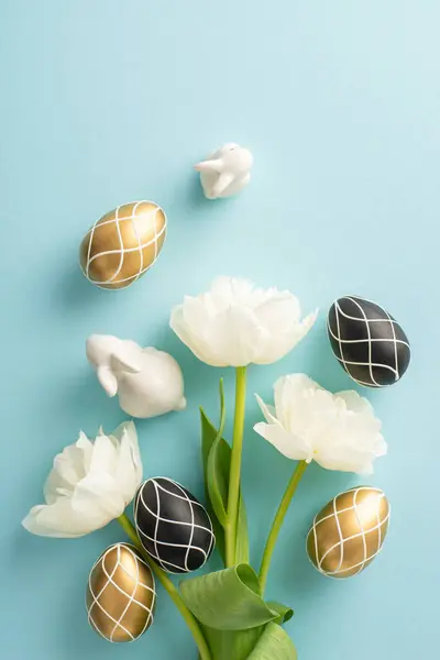 stock image Easter sophisticated setting. Top view snapshot featuring lavish black and gold eggs, bunny statuettes, bunch of delicate tulips, on a pale blue background with margin for messages