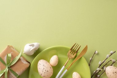 Charming Easter composition from top view, with natural gift wrapping, dining implements, bunny statuette, quail eggs, pussy willows laid out on pastel green background, including space for messaging