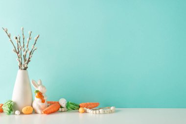 Easter display setup: Side view photo of a counter featuring a vase with pussy willow, a ceramic bunny figure, carrots for Easter bunny, assorted eggs, and beads on a pastel blue wall background