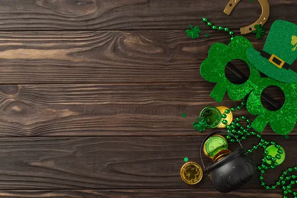 Delightful St. Patrick\'s composition seen from top view, consisting of clovers, pot with fortune coins, costume glasses, horseshoe, beads neatly placed on wood ground, leaving space for publicity