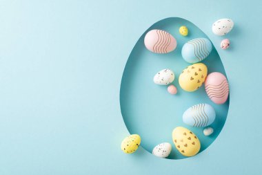 Paskalya bayramı yaratıcı sunumu. Renkli yumurtaların üst görüntüsü, açık mavi arkaplandaki yumurta şeklindeki bir açıklıktan görülebiliyor, selamlaşma ya da reklamlar için yer sunuyor.