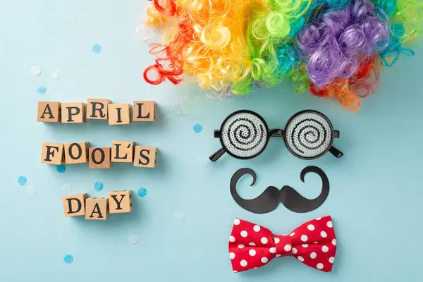 Jovial April Fool's setup: top view of wood letters spell 'April Fool Day', paired with a clown wig, mustache, bow tie, and eyeglasses, on a pastel blue background with scattered confetti