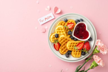 Anneler Günü brunch sahnesi: kalp waffle 'larının, çileklerin, yabanmersinlerinin, reçellerin, karanfillerin ve açık pembe bir arkaplanda içten gelen bir mesajın üst görüntüsü.