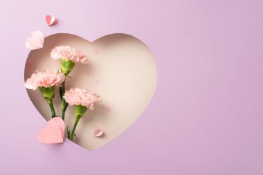 Anneler Günü cazibe teması. Pembe karanfillerin, dekoratif kalplerin ve konfetilerin üst görüntüsü, kalp şeklinde çerçevelenmiş, pastel bir eflatun arka planında, kişiselleştirilmiş mesajlar veya reklamlar için yer sağlıyor.