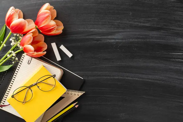 Extend Teacher\'s Day wishes with this setup. Overhead shot showcases teacher essentials, tulips, gypsophila, and chalk on a blackboard, providing space for text or promotional content
