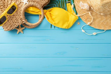 Yaz mevsiminin temel eşyalarından oluşan bir koleksiyon. Güneş gözlüğü, hasır şapka ve parlak mavi ahşap bir yüzeye dizilmiş sarı bir mayo. Yaz tatili ve plaj kaçamaklarının ruhunu somutlaştırıyor.