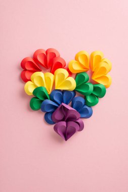 LGBTQ + gururunu sembolize etmek için gökkuşağı spektrumunda düzenlenmiş canlı dikey kağıt kalpler, yumuşak pembe bir arka planda sunulur, kabullenme ve aşk ruhunu yakalar.