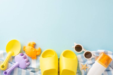 Çocukların plaj tatili fikri. Kum oyun setinin yukarıdan çekimleri: kürek, tırmık, deniz yaratığı kalıbı, SPF kremi, güneş gözlükleri, mini terlikler, plaj minderi, yumuşak mavi zeminde deniz yıldızı.