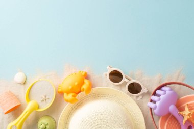 Çocukların yaz macerası teması. Plaj oyun setinin yukarıdan çekimleri: kova, kürek, yengeç kalıbı, hasır şapka, güneş gözlükleri, kum, kabuklar, yumuşak mavi arka planda yıldız balıkları, metin için yer var