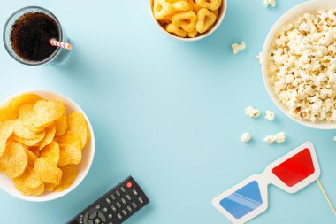 Ev yapımı bir sinema macerasına dalın: patlamış mısır, cips, soda ve uzaktan kumandanın üst görüntüsü. Pastel mavisi üzerine film temalı dekorasyonlar, metin veya reklam yerleştirme için ideal