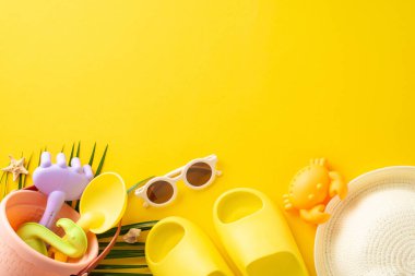Sarı arka planda duran bir kova, kürek ve güneş gözlüğü de dahil olmak üzere bir dizi yaz plaj oyuncağı güneşli bir plaj gününün özünü yakalıyor.
