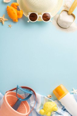 Çocuk dostu plaj tatili konsepti. Kum oyun topluluğunun dikey üst görüntüsü: kova, kürek, tırmık, deniz yaratığı kalıpları, SPF losyonu, hasır şapka, güneş gözlüğü, plaj havlusu, yumuşak mavi yüzeyde deniz yıldızı