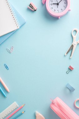 Renkli okul malzemelerinin dikey yaratıcı gösterisi. Not defterleri, kalemler ve pastel mavi bir yüzey üzerinde bir saat de dahil. Eğitici ve organizasyonel temaları temsil etmek için mükemmel