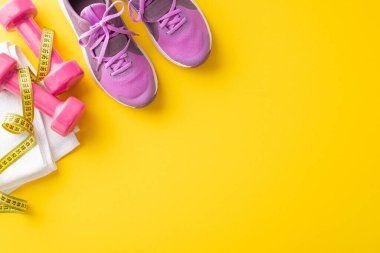 Pembe spor ayakkabılar, dambıllar, havlu ve canlı sarı bir arka planda ölçüm bandı, spor, spor ve sağlıklı bir yaşam tarzını temsil ediyor.