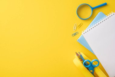 Not defterleri, makas, büyüteç ve ataç da dahil olmak üzere sarı arka plandaki parlak okul malzemelerinin en üst görüntüsü