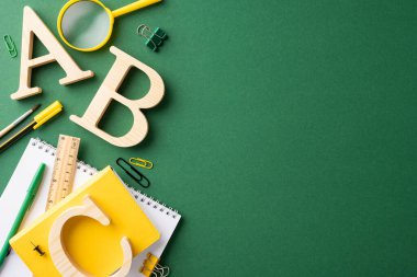 Okul gereçleri ve kırtasiye malzemelerinin en üst görüntüsü arasında defterler, kalemler, cetvel, büyüteç ve yeşil arka planda ahşap harfler yer alıyor.