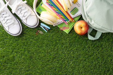 Back to school essentials including white shoes, colorful supplies, and an apple placed on green grass clipart