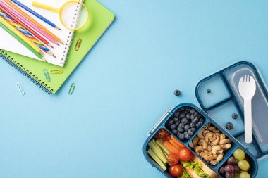 Notlar ve renkli kalemlerin yanında meyve, sebze ve fındıklı sağlıklı bir okul beslenme çantasının en üst görüntüsü.