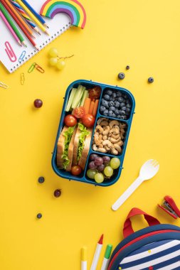 Değişik meyve, sebze ve sandviçlerle dolu renkli bir beslenme çantasında sağlıklı bir okul atıştırmasının dikey görüntüsü.