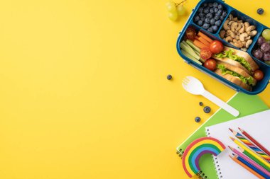 Sebzeler, meyveler, fındıklar ve parlak sarı arka planda renkli kalemlerle dolu bir defter içeren sağlıklı bir okul beslenme çantasının düz görüntüsü.