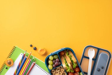 Sağlıklı bir okul atıştırması, beslenme çantası, defterler ve parlak sarı arka planda kalemler.