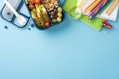 Renkli okul malzemelerinin yanı sıra sandviç, meyve ve sebzeli sağlıklı bir okul atıştırmalık kutusunun en üst görüntüsü.