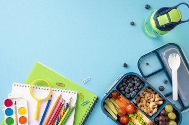 Mavi arka planda renkli kırtasiye malzemelerinin yanında taze meyve ve sebzelerle sağlıklı bir okul abur cuburunun üst görüntüsü.