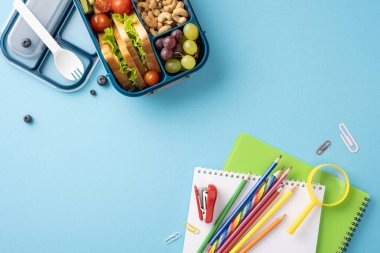 Sağlıklı bir okul beslenme çantasının en üst görüntüsü. Mavi arka planda renkli kırtasiye malzemelerinin yanında meyve ve atıştırmalıklar var.