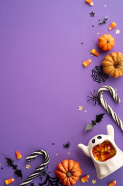 Dikey bir Cadılar Bayramı arka planında mor bir yüzey üzerinde şeker, balkabağı ve ürkütücü süslemeler yer alıyor.