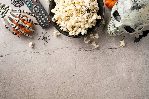 stock image Halloween-themed setup featuring a bowl of popcorn, remote control, skeleton hand, skull, and spider decorations on a concrete background