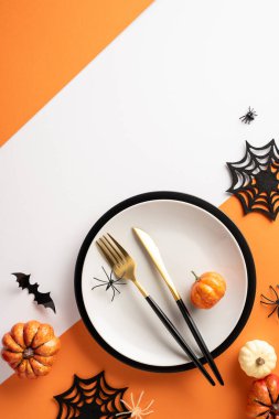 Beyaz tabak, altın çatal bıçak, minyatür balkabağı, örümcekler ve turuncu arka planda yarasa kesikleri içeren bayram dikey masa ayarı.