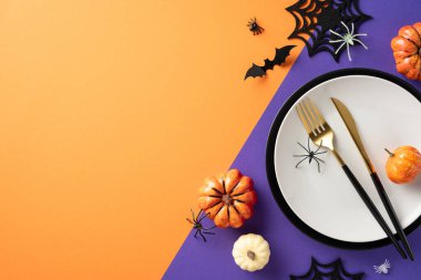 Cadılar Bayramı temalı masa ortamının üst görüntüsü. Kabaklar, yarasalar, örümcek ağları ve turuncu ve mor arka planda çatal bıçak takımı.