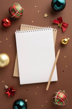 Kahverengi arkaplanda renkli Noel süsleriyle çevrili boş bir not defterinin üst dikey görüntüsü