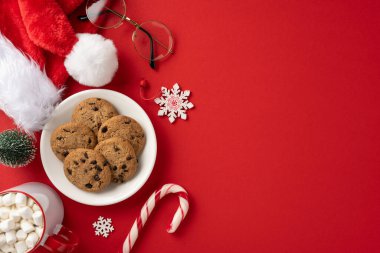 Bir tabak kurabiye, şeker kamışı ve Noel süslemelerinin yer aldığı bir tatil sahnesi canlı kırmızı arka planda, Noel Baba 'yı temsil ediyor.