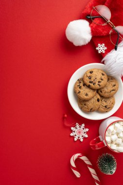 Kurabiyeler, Noel Baba şapkası, şeker kamışı, marşmelov ve canlı kırmızı arka planda kar taneleriyle dolu rahat bir Noel ortamının dikey görüntüsü.