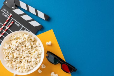 Patlamış mısır kasesinin üst görüntüsü, alkış tahtası ve renkli arka planda 3D gözlükler, eğlenceli bir film gecesi atmosferini temsil ediyor.