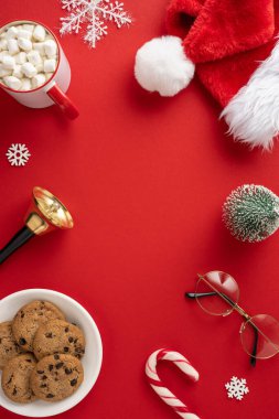 Dikey tatil sahnesinde Noel Baba şapkası, kremalı sıcak çikolata, kurabiye ve kırmızı arka planda şenlikli süslemeler var.