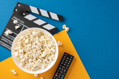 Bir kase patlamış mısır, film seti ve uzaktan kumandalı mavi ve sarı arka plan, bir film gecesi deneyimini temsil ediyor.