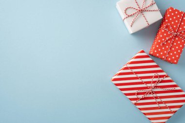 Kırmızı ve beyaz tasarımlı güzel paketlenmiş üç Noel hediyesi açık mavi bir yüzeyde düzenlenerek neşeli ve neşeli bir atmosfer yaratılıyor.