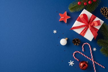 Güzel paketlenmiş, kırmızı kurdeleli, mavi arka planda tatil süslemeleriyle çevrili bir Noel hediyesi. Şeker kamışları, süsler ve çam dalları içerir