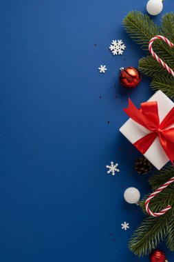 Kırmızı kurdeleli bir Noel hediyesi. Çam dalları, baston şekerler ve mavi arka plandaki süsler gibi neşeli elementlerle çevrili.