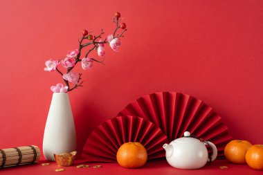 Mandalina, sakura çiçekleri ve kırmızı vantilatörlerin bulunduğu Çin Yeni Yıl 2025 elementlerinin canlı kırmızı arka planda sergilenmesi, şans ve refahı simgeliyor.