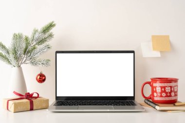 Dizüstü bilgisayarı, şenlikli süslemeleri ve İskandinav tarzı minimalist çalışma alanı. Kırmızı bir fincan, hediye kutusu ve küçük bir Noel süsü. Sıcak bir kış ortamı yaratıyor.